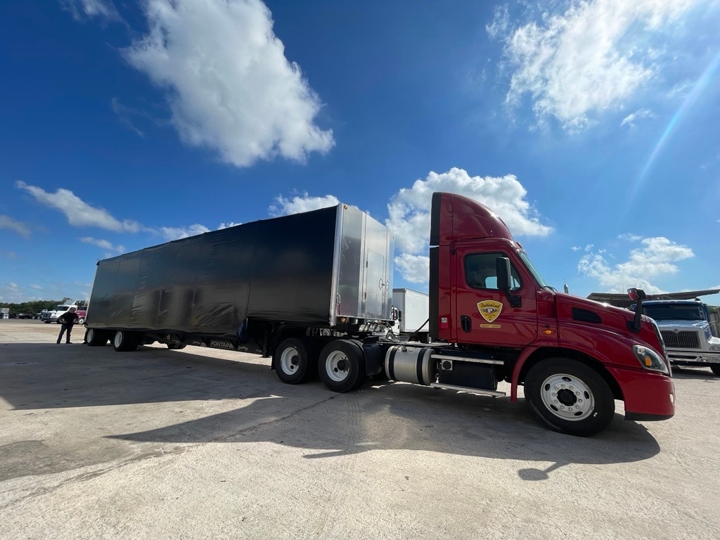 Palletized red Cascadia pulling a black conestoga flatbed trailer