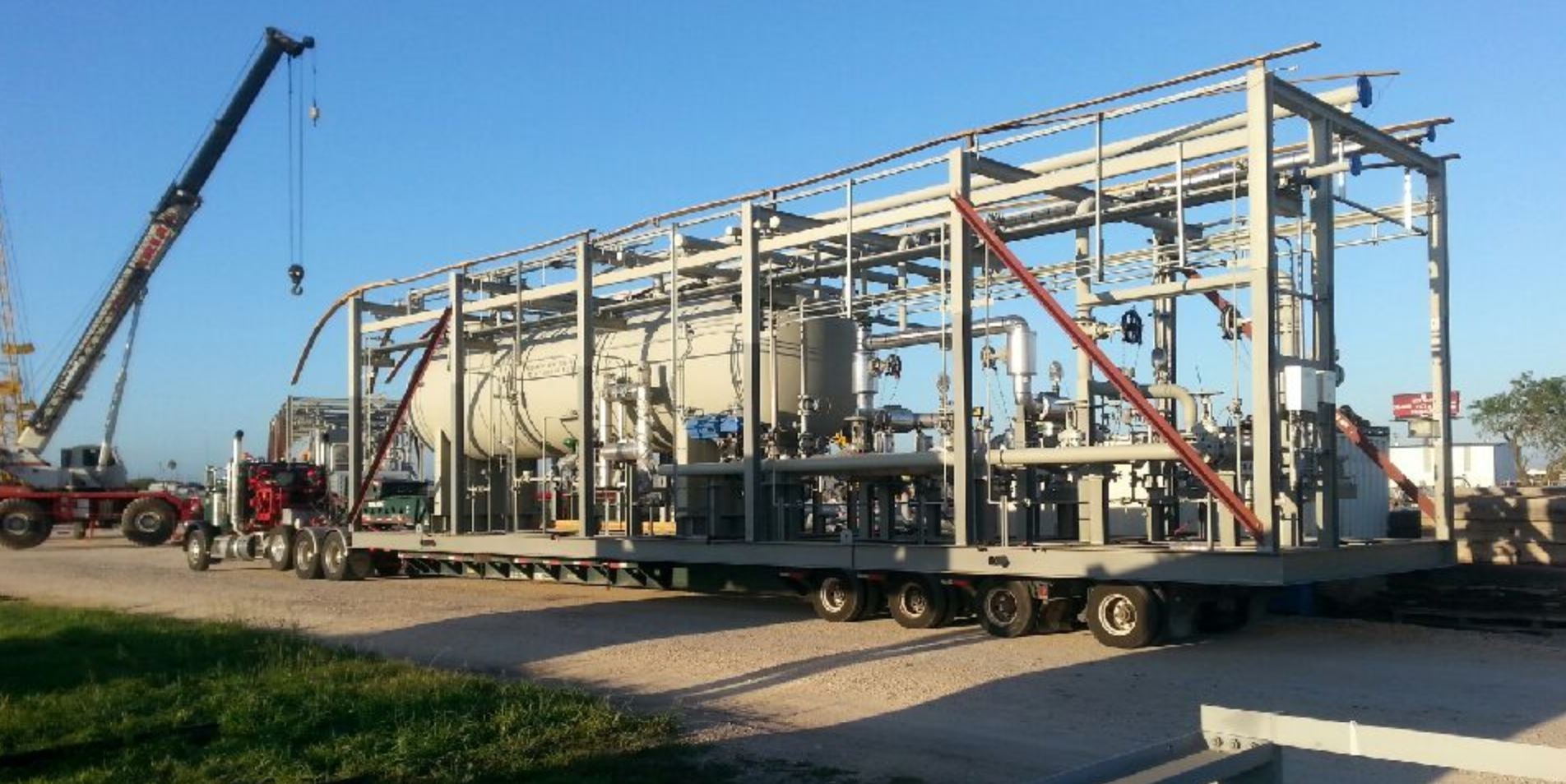 Palletized truck hauling large modular processing unit being crane-loaded at industrial yard