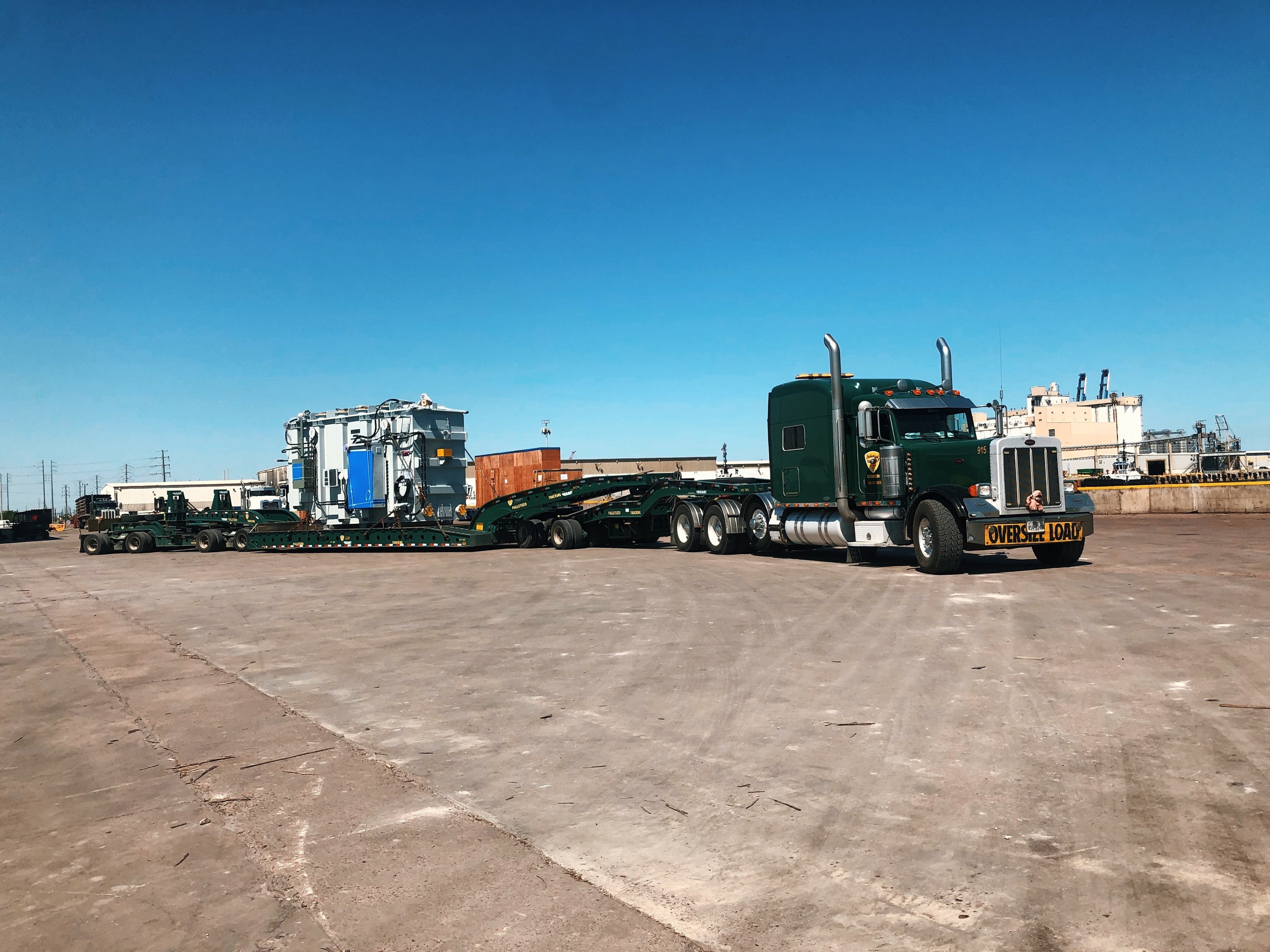 Palletized green Peterbilt hauling oversized transformer on multi-axle lowboy at port